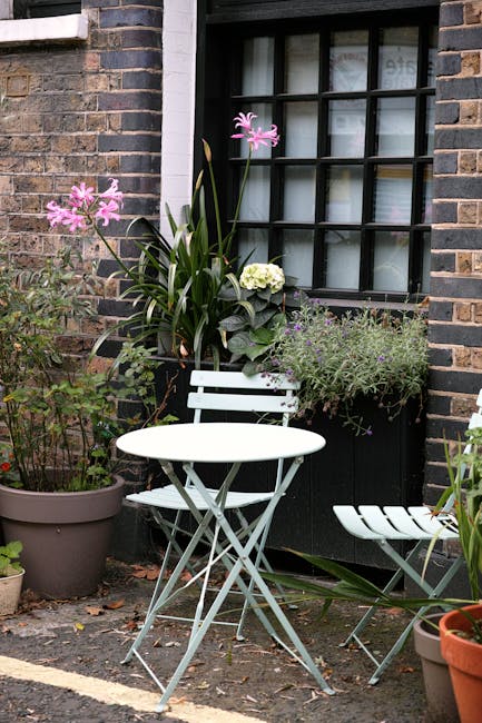 A cozy front garden scene featuring a small, white metal bistro table with matching chairs positioned against a brick wall exterior of a house. The window above has black grid panes and a white frame.