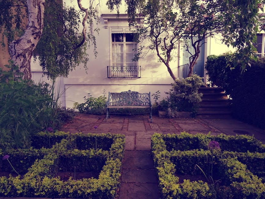 A cozy front garden scene featuring a small, white metal bistro table with matching chairs positioned against a brick wall exterior of a house. The window above has black grid panes and a white frame.