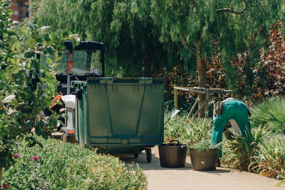 A gardener wearing teal work clothes is bent over, tending to plants in a lush, well-maintained garden. They are surrounded by various green plants, including leafy shrubs and tall, spiky foliage, wit