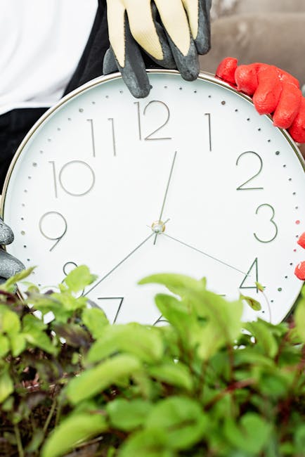 A gardener wearing teal work clothes is bent over, tending to plants in a lush, well-maintained garden. They are surrounded by various green plants, including leafy shrubs and tall, spiky foliage, wit